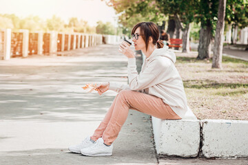 Woman in white hoodie with paper cup of coffee is sitting outside in autumn park.