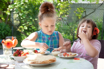 
Sibling sister children have a fun breakfast at their grandmother's cottage. Positive smiling kids, summer breakfast. Pancakes, strawberries, cherries, poppies