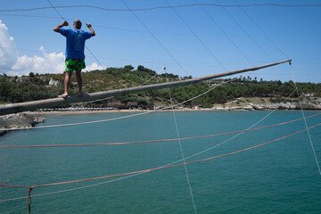 Naklejka premium Unrecognizable fisherman walks over a wooden pole and holds onto the ropes of a traditional fishing trabucco at the beach of Vieste, Italy