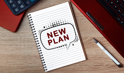 Notebook with text NEW PLAN. Notebook on desk with laptop and a pen.