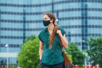 A woman in a medical face mask walks thrusting a hand into a pocket of trousers, holds a bag in downtown. Girl keeping social distance wears a protective face mask to avoid the spread of coronavirus