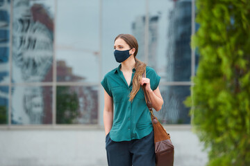 Cute woman in medical face mask walks thrusting hand into a pocket of trousers, holds a bag in downtown. Girl keeping social distance wears a protective face mask to avoid the spread of coronavirus