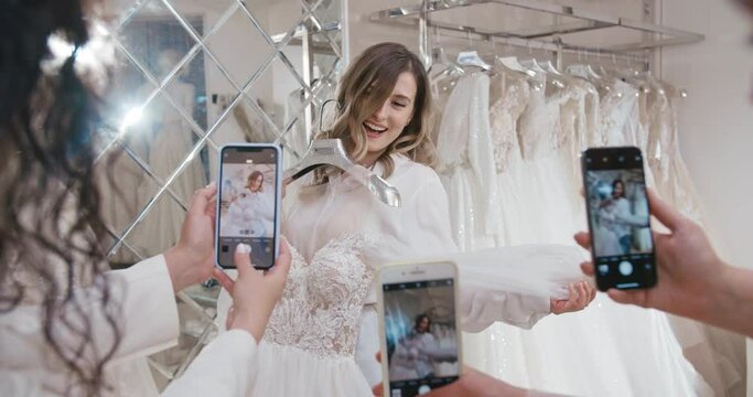 Three bridesmaids taking pictures of beautiful blonde girl with chosen wedding dress using phones. Bride posing for photo with dress on hanger in front of mirror in wedding salon.