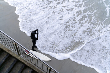 Surfer wearing wetsuit with surfboard watching ocean waves crash over rocks at St. Clair beach,...
