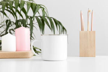 Enamel mug mockup with workspace accessories and candles on a white table and a palm plant.