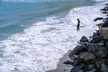 Surfer wearing wetsuit with surfboard watching ocean waves crash over rocks at St. Clair beach, Dunedin, New Zealand.