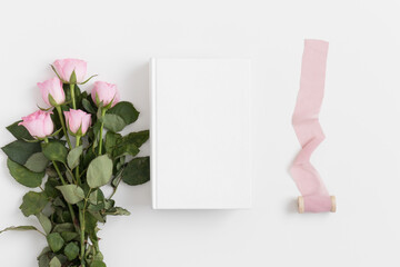 White book mockup with a bouquet of pink roses and a silk ribbon on a white table.