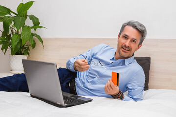 The man with gray hair lying on the bed with a laptop and holding eyeglasses and banking card in your hands, looking and smiling at the camera.