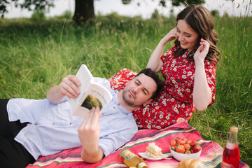 Young couple sits on planket outdoork and read book on mini picnic. Man lies on her knees