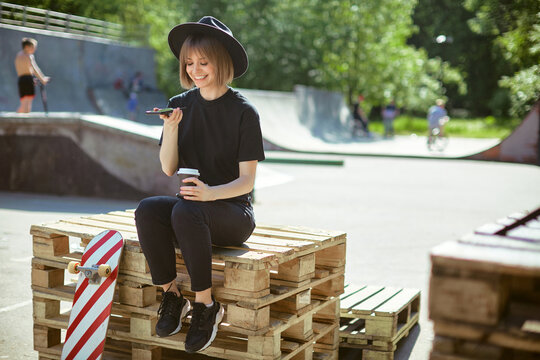 Smiling girl chat on phone sitting on wooden pallet in park with skateboard and cup coffee to go, photo for ad or blog
