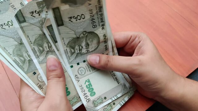 Hands counting new Rs 500 Rupees Indian Currency notes against a wooden bokeh background. A torn note is found in the bundle and separated