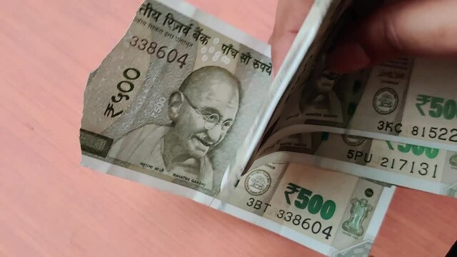 Hands counting new Rs 500 Rupees Indian Currency notes against a wooden bokeh background. A torn note is found in the bundle and separated