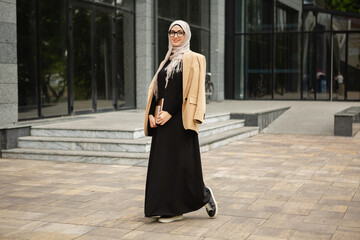 modern stylish muslim woman in hijab, business style jacket and black abaya walking in city street...