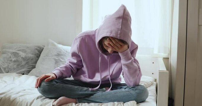 Full length frustrated teenage girl sitting on bed, feeling stressed alone at home. Depressed adolescent schoolgirl suffering from puberty psychological problem, having headache, thinking of suicide.