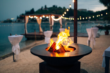 Grill with flames inside. Round table-cooking surface. On the beach, in the background of the gazebo by the water with garlands, in the twilight light. - obrazy, fototapety, plakaty