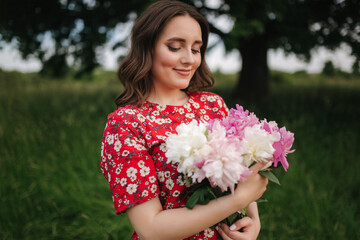 Fototapeta premium Portrait of Attractive young woman in red dress with beautiful flowers walking in the field in front of big tree