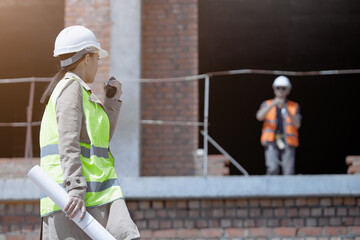 An engineer girl with a walkie in her hands on a construction site in a protective vest and hard hat, a worker in the background