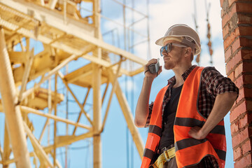 A construction site worker in a protective vest and hard hat communicates on a walkie talkie against the background of a construction crane