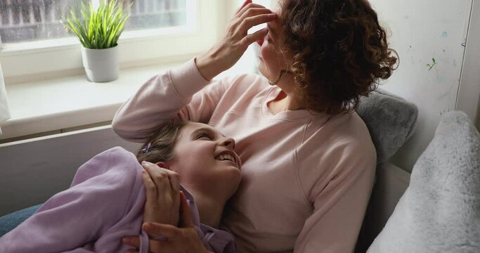 Smiling woman lying on comfortable bed or couch, cuddling laughing adolescent kid daughter. Happy teen girl having fun joking with affectionate mother, enjoying spending free time together at home.