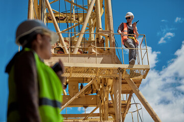 Girls engineer with radio in hand on a construction site in a protective vest and hard hat, in the background a worker on a yellow crane. Selective focus
