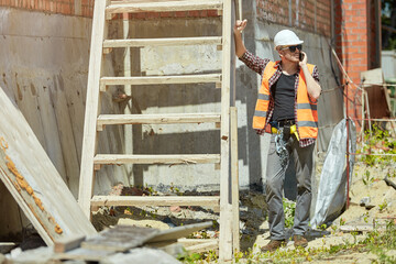 Construction site worker in a protective vest and hard hat, resting and using a mobile phone