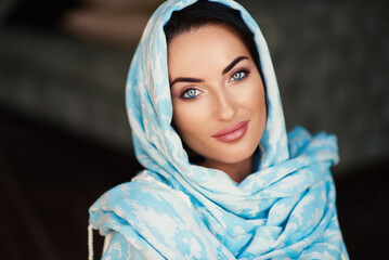 Fototapeta premium Young woman of oriental appearance in a blue scarf. Beauty portrait of arabian or indian girl with perfect makeup and handmade accessories