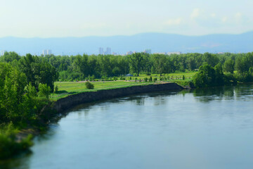 Obraz premium Sheer bank of river, water, green meadow, forest. Away the city and mountains. Concept of a layered landscape in summer. Tilt-shift effect. Selective focus.