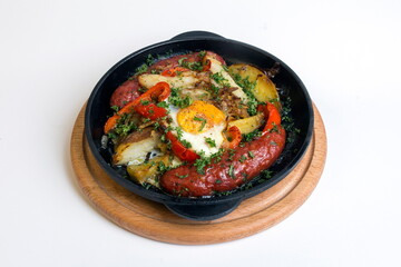 Dish on a black pan, which stands on a wooden round stand. Baked potatoes with sausages and red pepper. Sprinkled with green parsley.