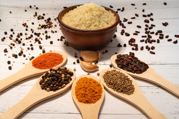 Ingredients for cooking pilaf: rice in a ceramic bowl in the background, garlic in the foreground, barberry, cumin, saffron and red pepper in wooden spoons and scattered on the table.