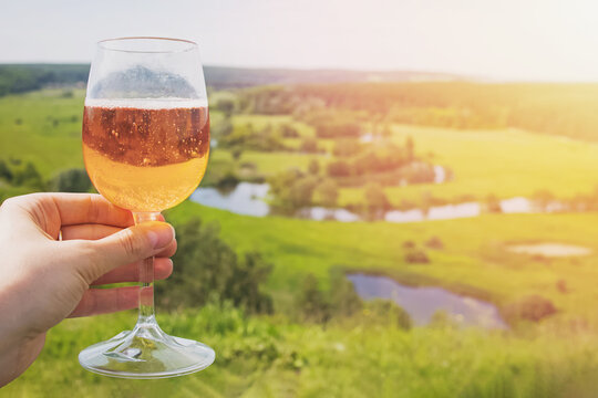Hand holding a glaa of sider or other alcoholic sparkling drink with the view to the beautiful green meadow in countryside
