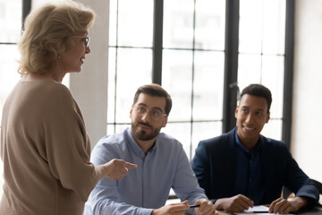 Diverse colleagues listening to mature business coach at briefing, middle aged businesswoman mentor wearing glasses speaking, explaining strategy, training staff at corporate meeting in boardroom