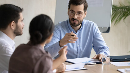 Confident businessman wearing glasses discussing project strategy or opportunities with diverse business partners at meeting, sitting at table in boardroom, leader giving instructions to employees