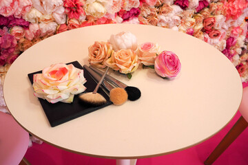 closed makeup palette brushes and artificial roses on a round pink table against a wall of flowers . cosmetic
