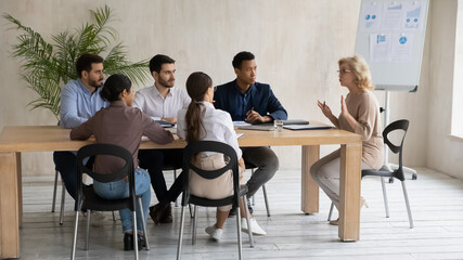 Middle aged businesswoman wearing glasses, team leader holding corporate meeting, diverse employees team sitting at table in boardroom, listening to executive, mentor coach giving instructions