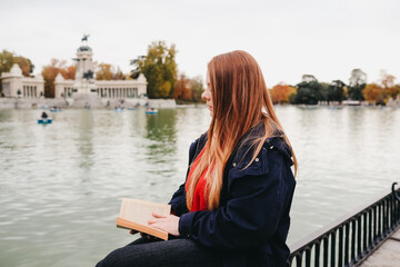 atractiva mujer joven pelirroja con pecas sentada en un banco leyendo y en parque pensativa delante...