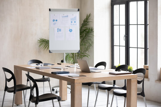 Modern office boardroom with flip chart with statistics and diagrams and conference table with laptop and documents for corporate briefing or group negotiations, empty meeting room interior