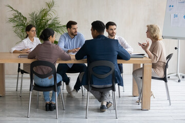 Serious middle aged businesswoman leading corporate meeting with diverse employees team, business people listening to mentor coach at briefing, sitting at conference table in boardroom