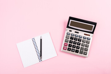 Large silver calculator with black pen and white paper notebook laying on a pink background. Concept of finance, business, calculations, education, school, math, accounting, computing, profit, tax