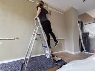 woman stands  on a ledder and paints the wall brown or taupe. Brown colour. Renovate a room -...