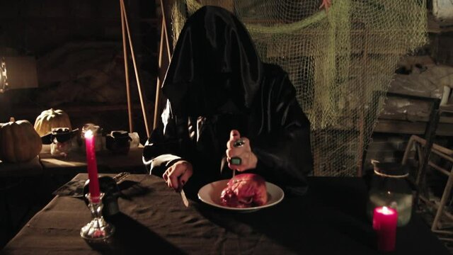 An evil dark sorcerer in a black mantle with a hood eats a raw heart at a table with a node and a fork. Raw meat. Dark magic with amulets, candles and artifacts