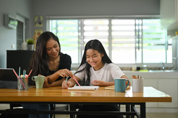 A mother is sitting with her daughter and teaching her homework at the wooden working desk.