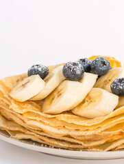 A dessert on a white plate topped with fruit. Thin pancakes with blueberry and banana