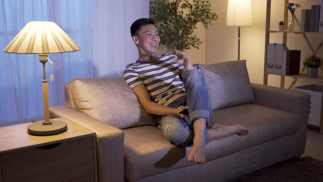 japanese guy spends leisure time watching funny dramas to release stress. relaxing non-working night asian male burst out laughter while hitting lap and pointing screen to make comment to the jokes