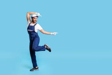 Full length funny joyful handyman in uniform standing in dance move and pointing copy space,...