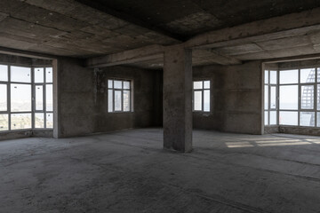 Large spacious room, illuminated by natural light from windows. Unfinished building interior. Repairs in the apartment. Empty dark abstract concrete interior.