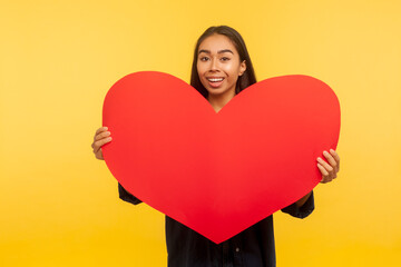 Portrait of beautiful happy girl with charming smile holding big read heart and smiling to camera, expressing love, greeting on Valentine's day. indoor studio shot isolated on yellow background