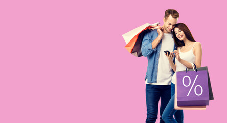 Holiday sales, store mall, retail, consumer concept - happy couple with shopping bags, looking at mobile phone, standing close to each other. Over rose pink color background.