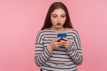 Portrait of serious attentive young woman typing on cell phone, reading blog, chatting in social network, using mobile app for online communication. indoor studio shot isolated on pink background