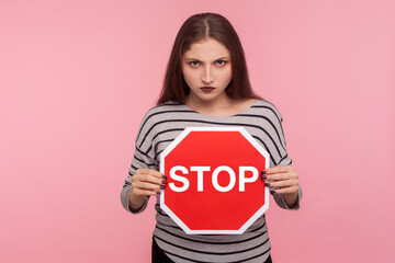 Way prohibited! Portrait of angry strict bossy girl in striped sweatshirt holding red Stop symbol, road traffic sign as warning of ban, forbidden access. indoor studio shot isolated on pink background