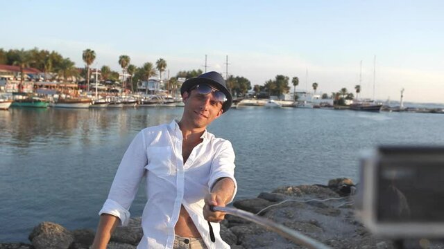 Young man wearing white shirt and hat posing for selfie with handstick while on vacation in exotic place on Mediterranean Sea. Man showing "V" for "victory" sign with his fingers. Holidays concept.